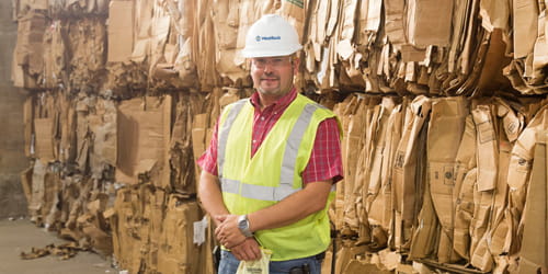 Um homem de colete e capacete em pé diante de papelão de fibra reciclado.